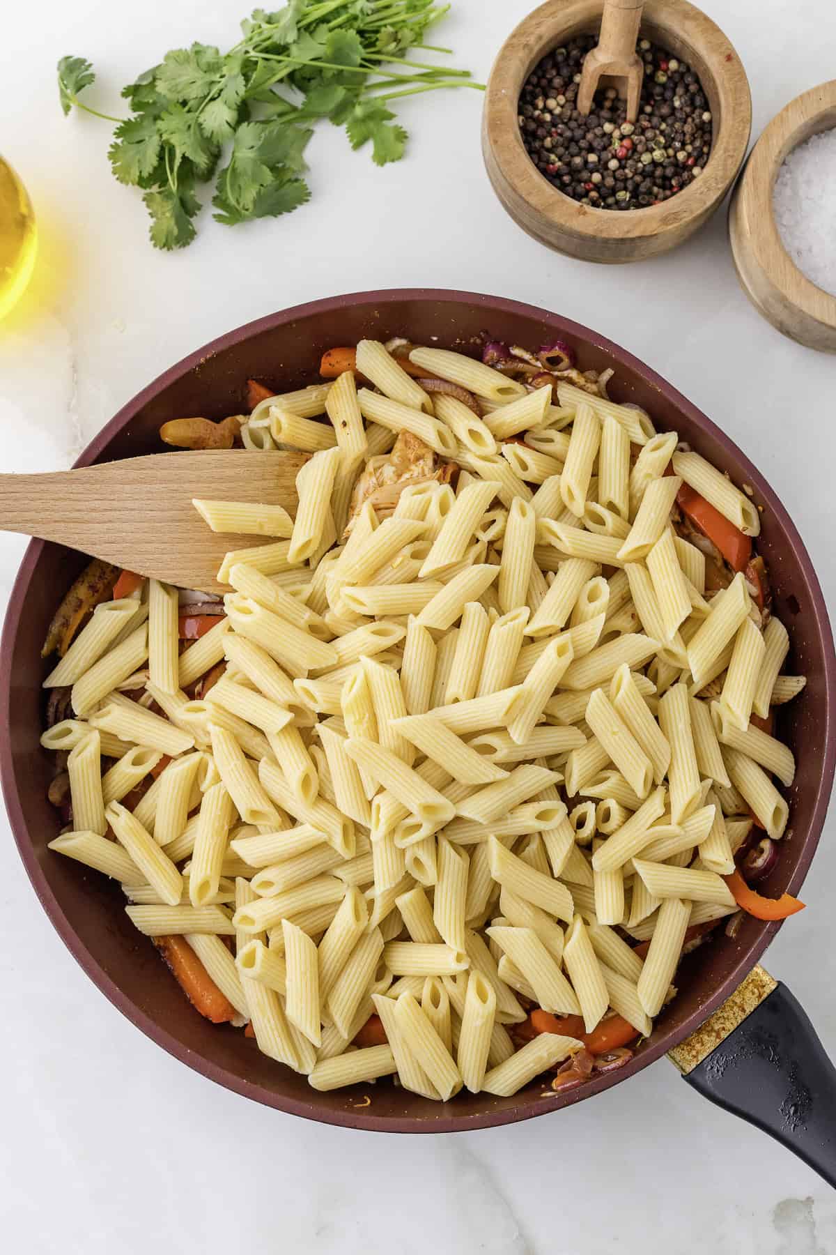 Penne pasta in a red skillet with vegetables and herbs, a wooden spatula in the mix. Salt and pepper in wooden bowls, cilantro and olive oil are nearby on a white surface.
