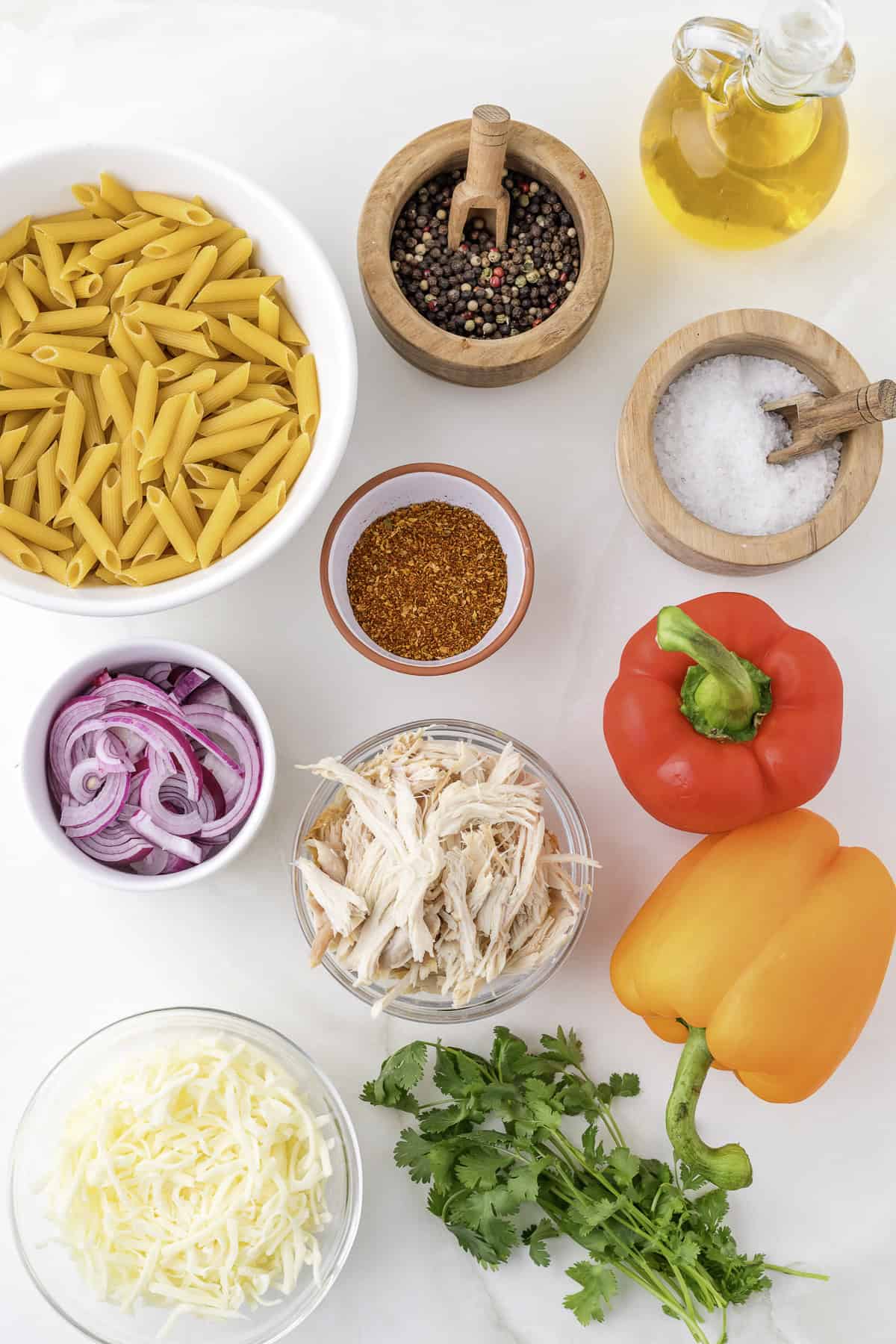 Ingredients on a white surface: penne pasta, shredded chicken, sliced red onion, shredded cheese, red and orange bell peppers, mixed peppers, salt, oil, spices, and cilantro.