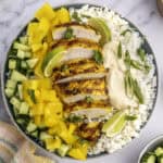 A bowl with sliced grilled chicken, yellow bell peppers, cucumber, lime wedges, rice, feta cheese, and hummus, garnished with herbs.