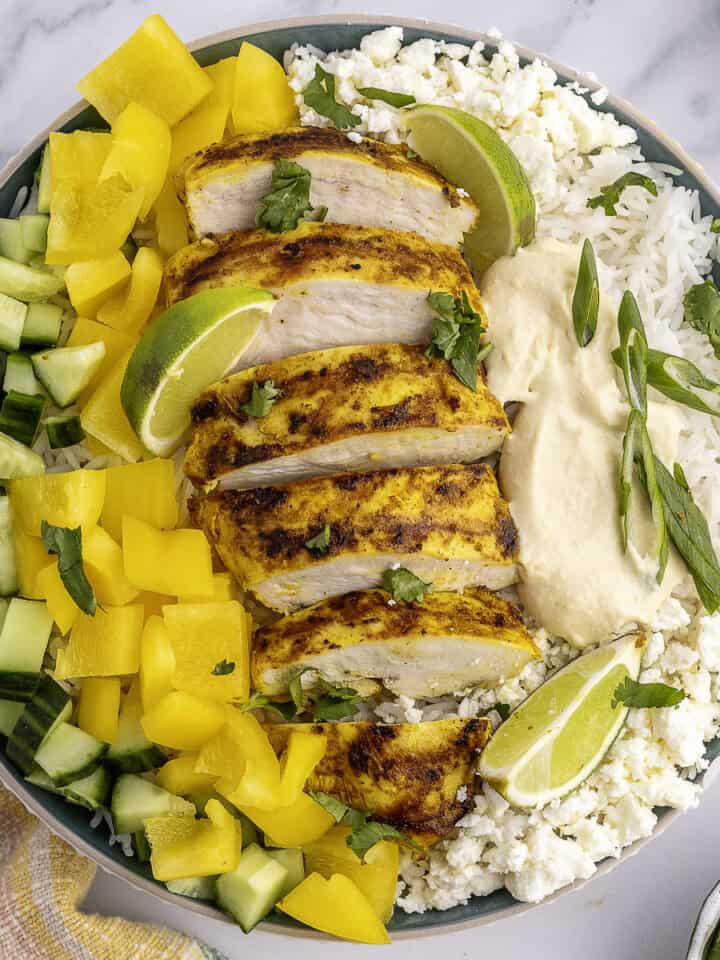 A bowl with sliced grilled chicken, yellow bell peppers, cucumber, lime wedges, rice, feta cheese, and hummus, garnished with herbs.
