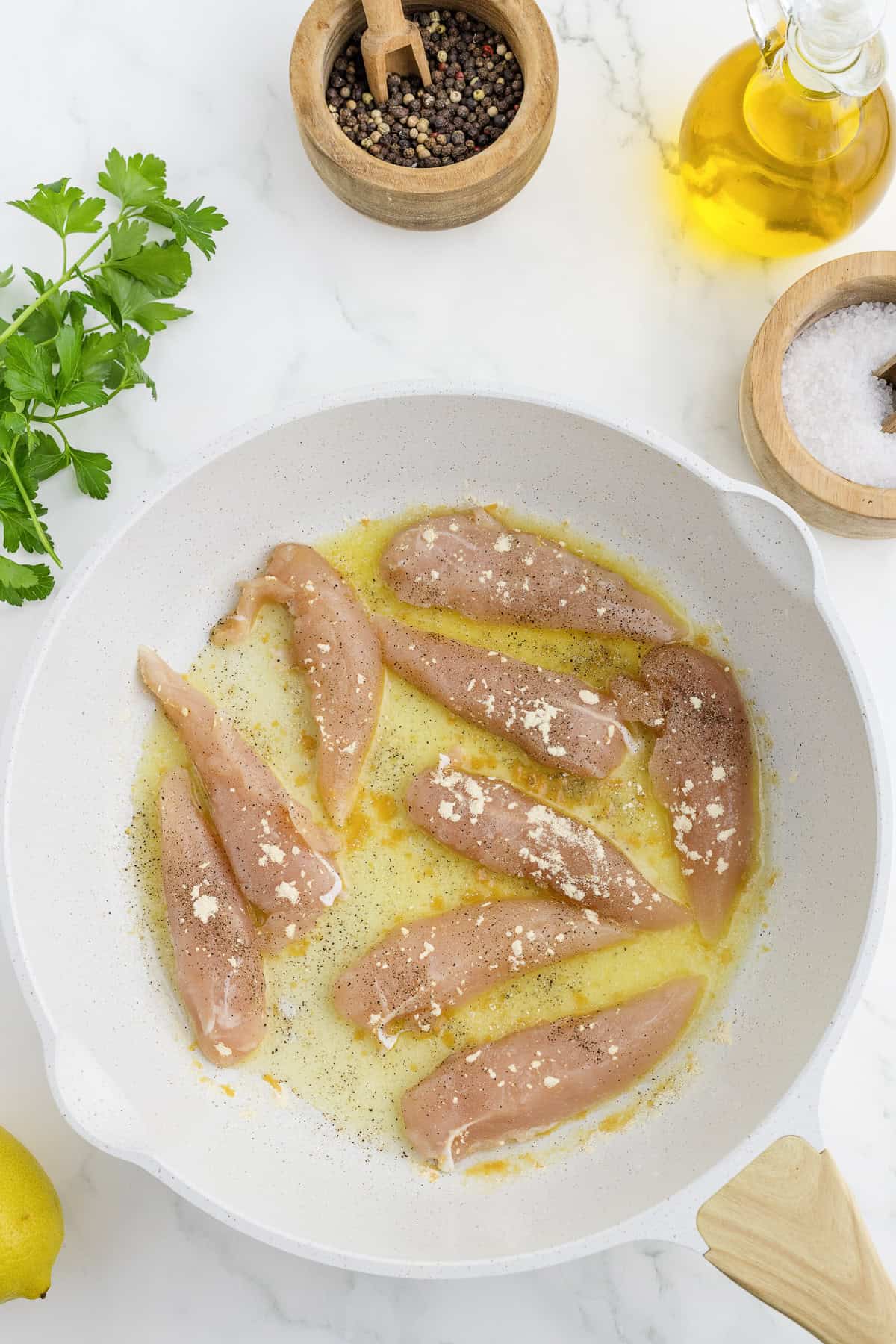 Raw chicken fillets are seasoned with salt and pepper in a white frying pan. Nearby are bowls of peppercorns and salt, olive oil, and fresh parsley.