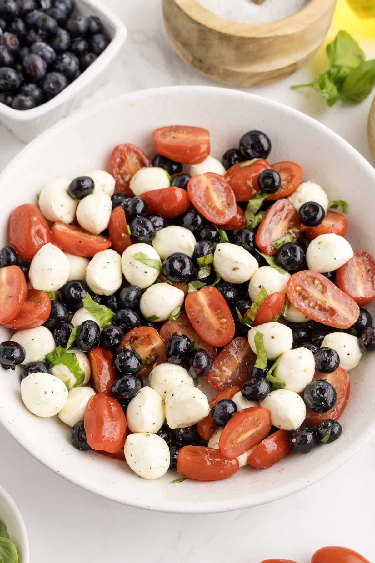 A white bowl filled with cherry tomatoes, blueberries, mozzarella balls, and basil leaves, lightly seasoned, sits on a white surface next to small bowls of salt and blueberries.