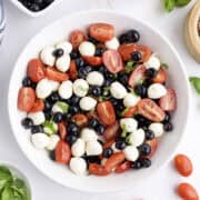 A white bowl filled with grape tomatoes, blueberries, mozzarella balls, and fresh basil leaves on a white surface.
