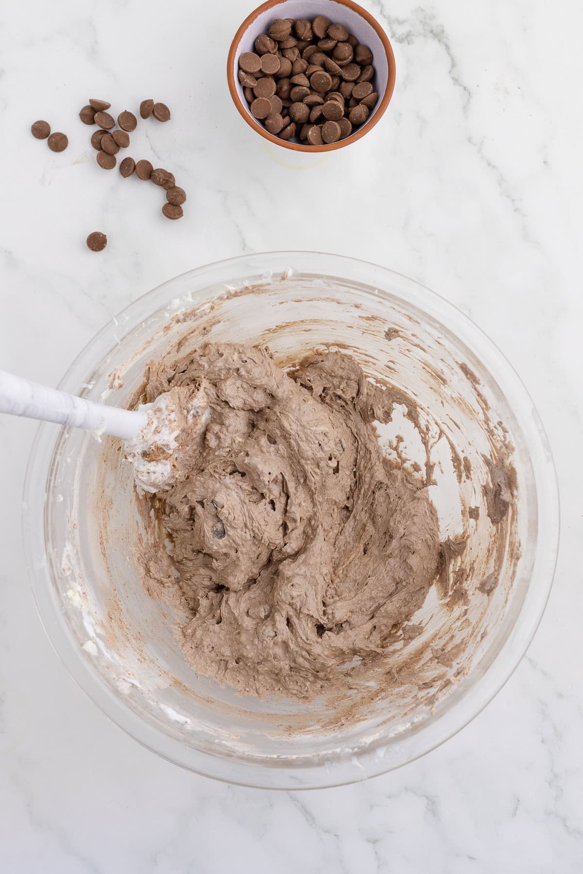 Bowl with chocolate chip cookie dough and a spatula on a marble surface. A small bowl of chocolate chips and a few scattered chips are in the background.