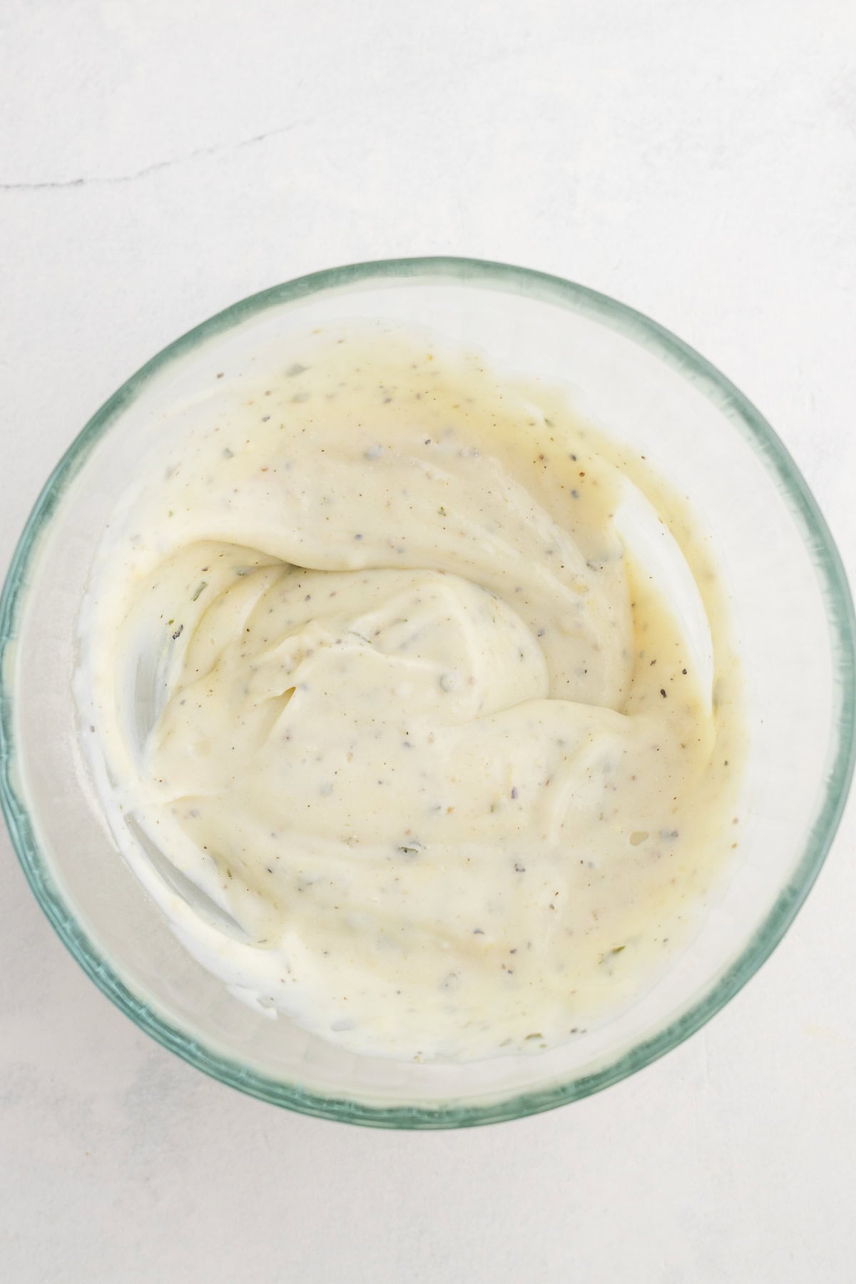 A glass bowl containing a creamy, white sauce with visible specks of herbs and spices on a light surface.