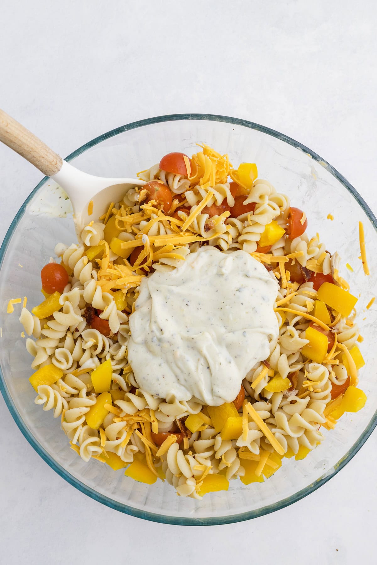 A glass bowl with rotini pasta, chopped yellow bell peppers, halved cherry tomatoes, shredded cheese, and a dollop of creamy dressing, with a white spoon resting inside.
