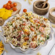 A bowl of creamy pasta salad with rotini, cherry tomatoes, bacon, shredded chicken, yellow bell peppers, and parsley, surrounded by ingredients on a white surface.