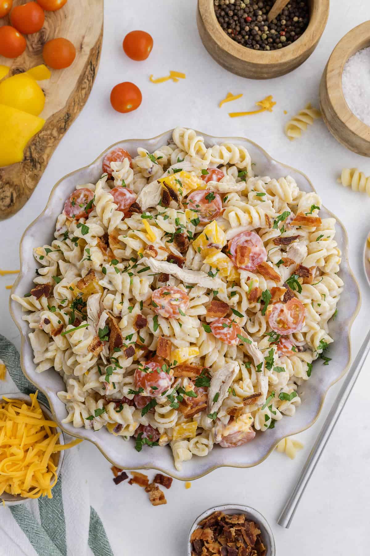 A bowl of rotini pasta salad with shredded chicken, cherry tomatoes, cheddar cheese, bacon, and chopped parsley on a white surface with ingredients nearby.
