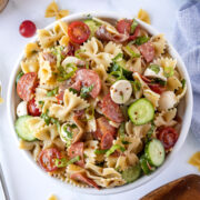 A bowl of pasta salad with bow-tie pasta, sliced pepperoni, cherry tomatoes, cucumber, mozzarella balls, and fresh herbs.