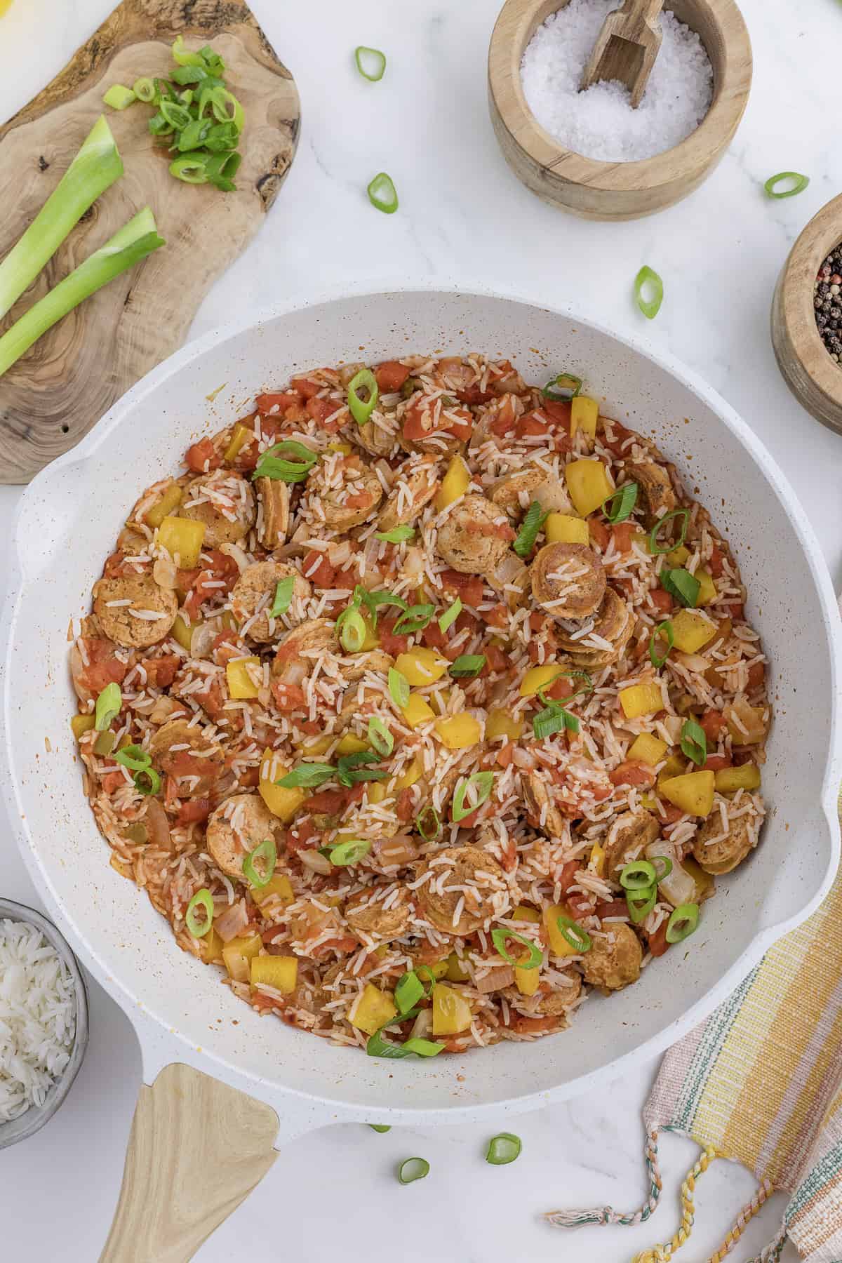 A skillet dish with sliced chicken sausage, diced tomatoes, yellow bell peppers, rice, and green onions. Wooden and ceramic bowls with ingredients surround the skillet on a marble surface.