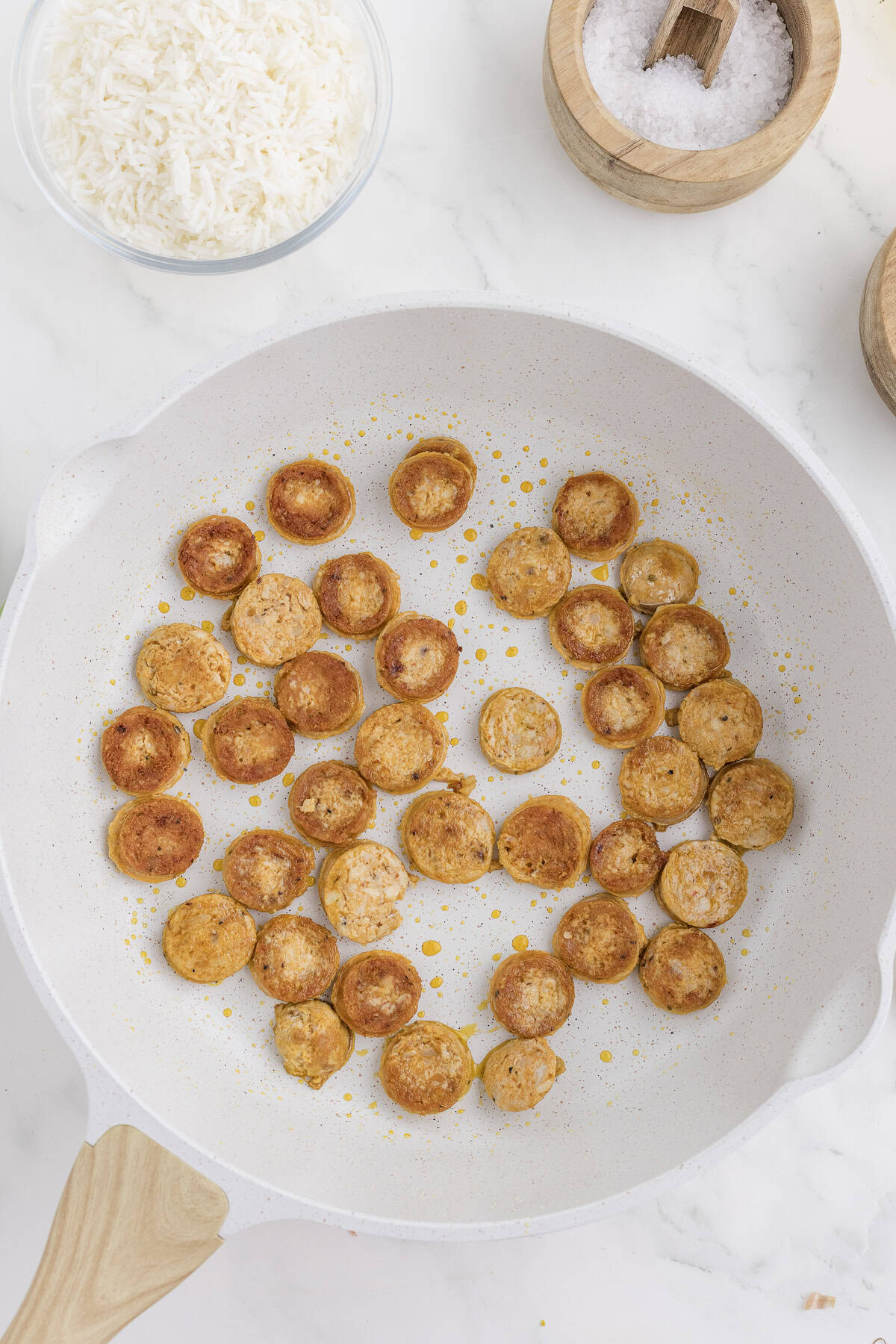 Sliced sausage pieces frying in a white pan with oil spots, alongside a bowl of white rice and a bowl of salt on a marble surface.