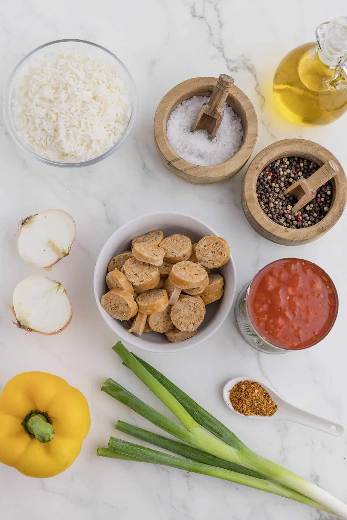 Various cooking ingredients including bell pepper, sausage slices, rice, spices, oil, salt, peppercorns, chopped onion, green onions, and tomato sauce arranged on a marble surface.