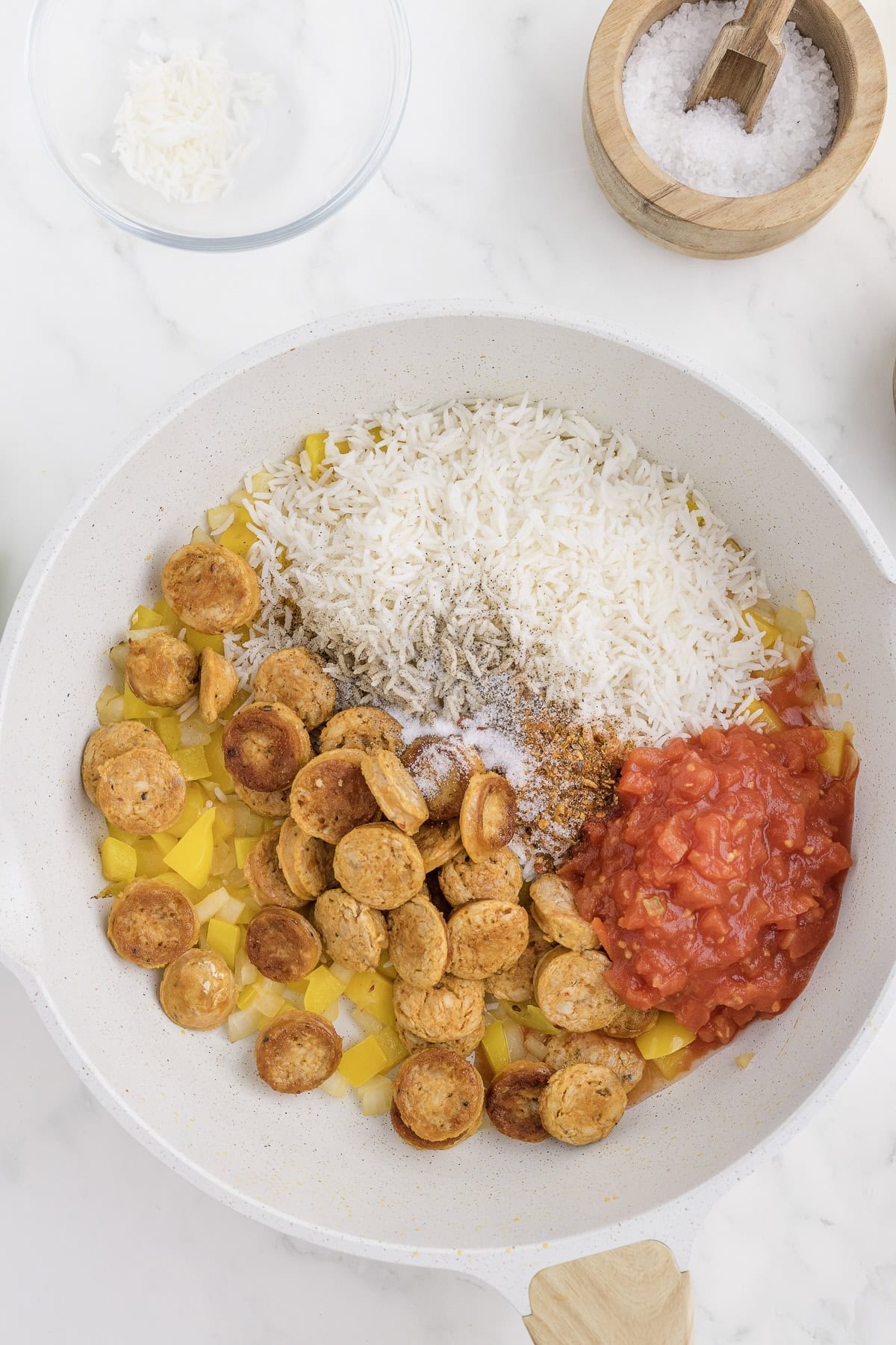 A skillet with sliced sausage, yellow bell peppers, uncooked rice, diced tomatoes, and seasoning. Small bowls of salt and shredded coconut nearby on a marble surface.