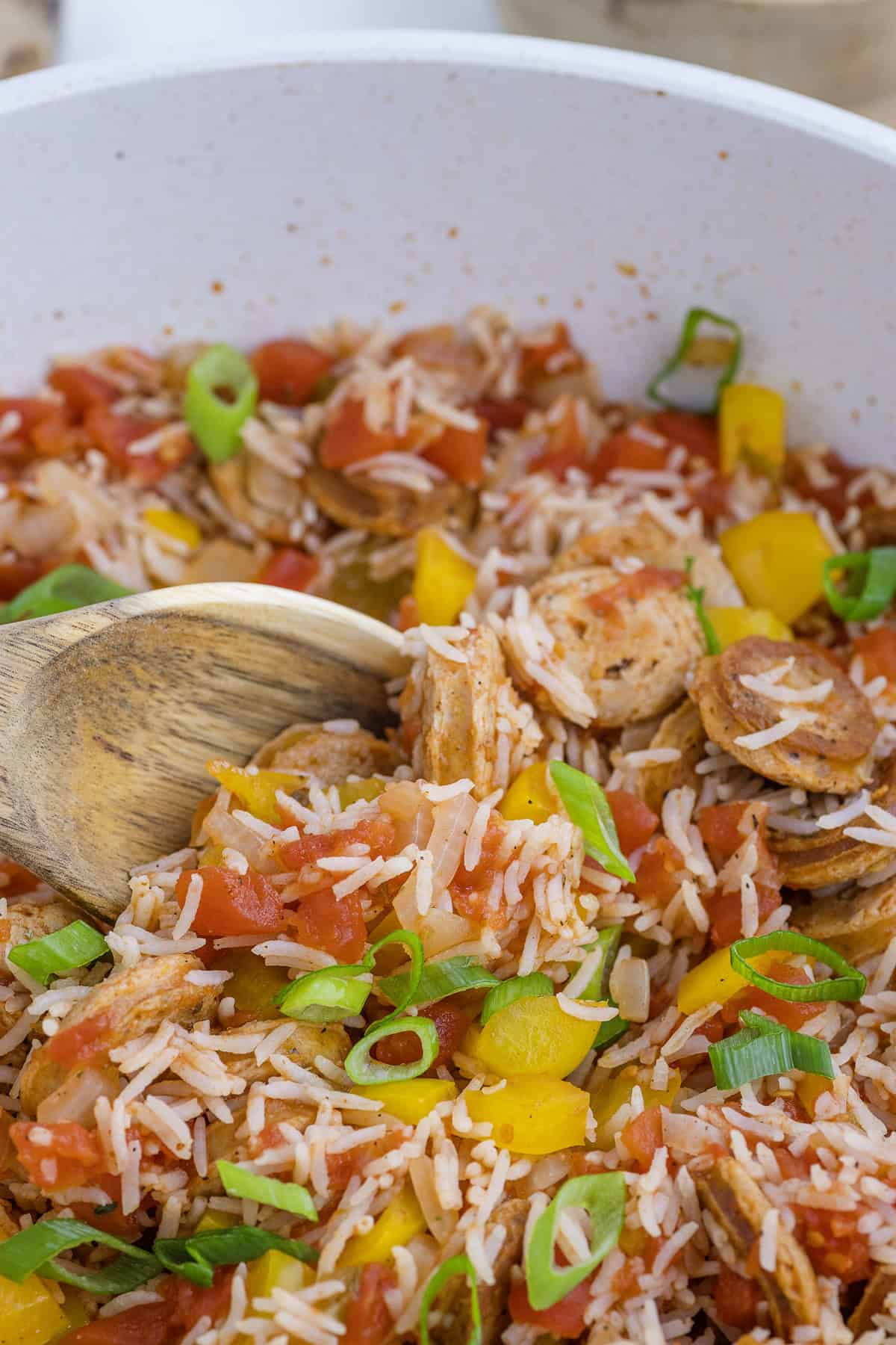 A pan filled with cooked rice, chopped sausages, diced tomatoes, yellow bell peppers, and green onions, with a wooden spoon stirring the mixture.