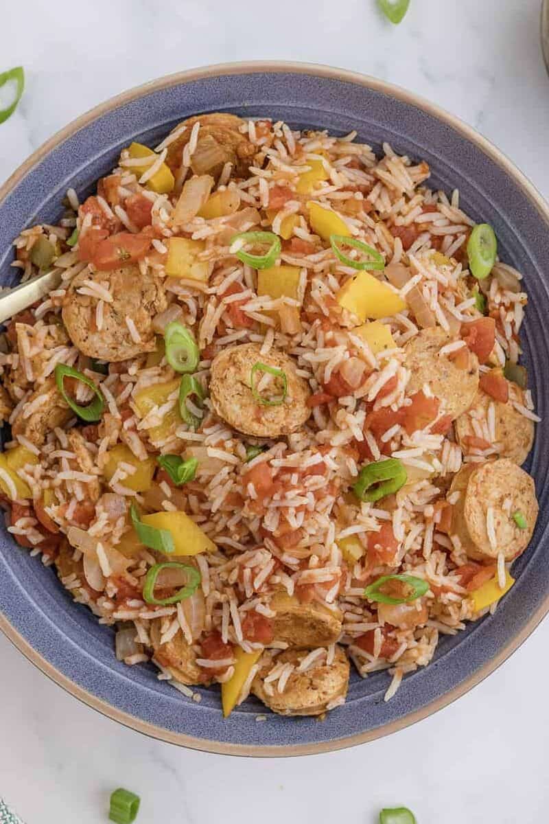 A bowl of rice mixed with sliced sausage, tomato, and yellow bell pepper, garnished with chopped green onions. It is surrounded by bowls of salt, pepper, and a bottle of oil on a marble surface.