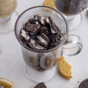 Clear glass mug filled with chocolate Oreo mug cake topped with crushed cookies. Fragments of cookies scattered on the white surface around the mug.