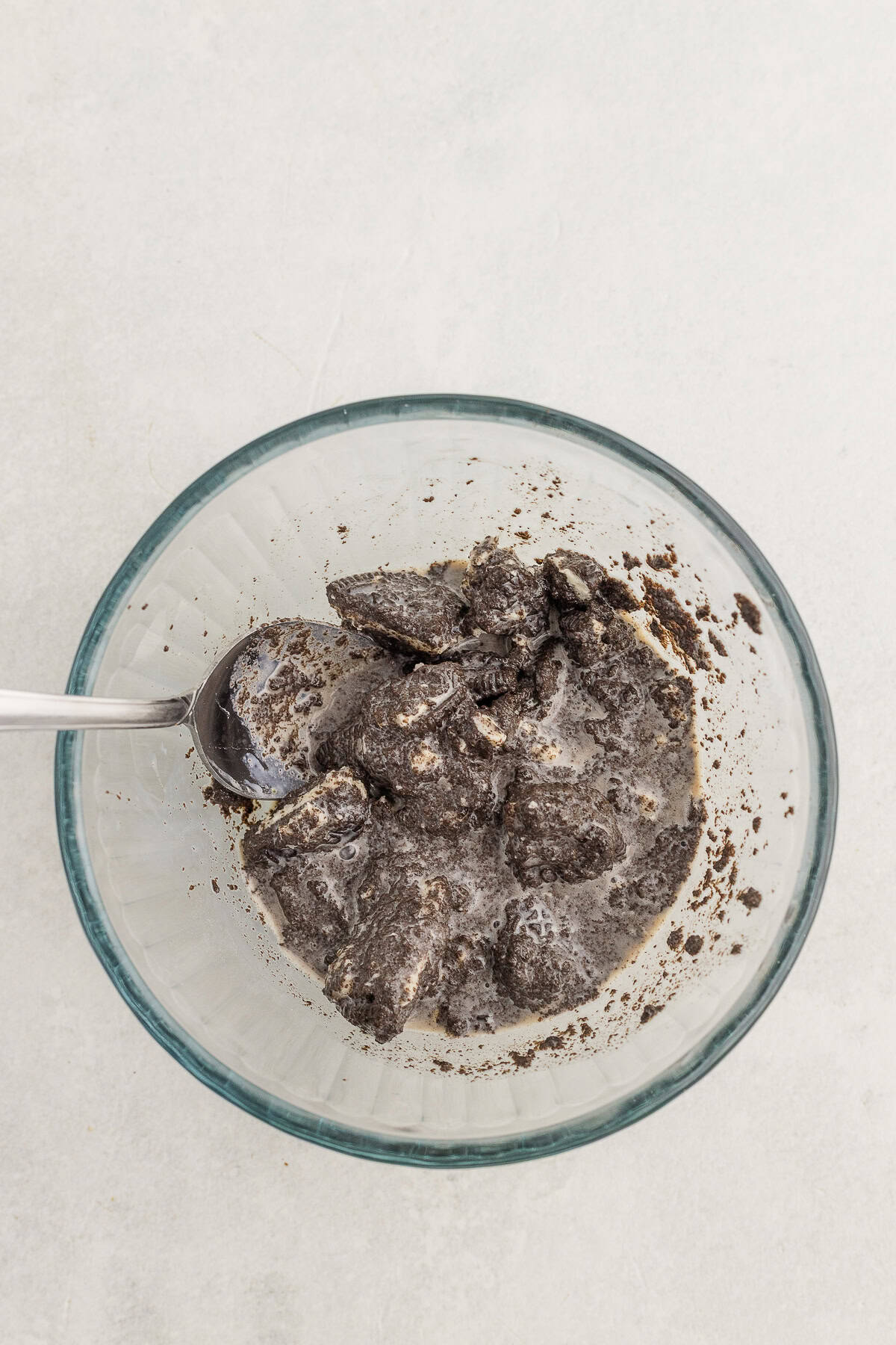 A glass bowl filled with crumbled chocolate cookies mixed with milk, and a spoon resting inside.