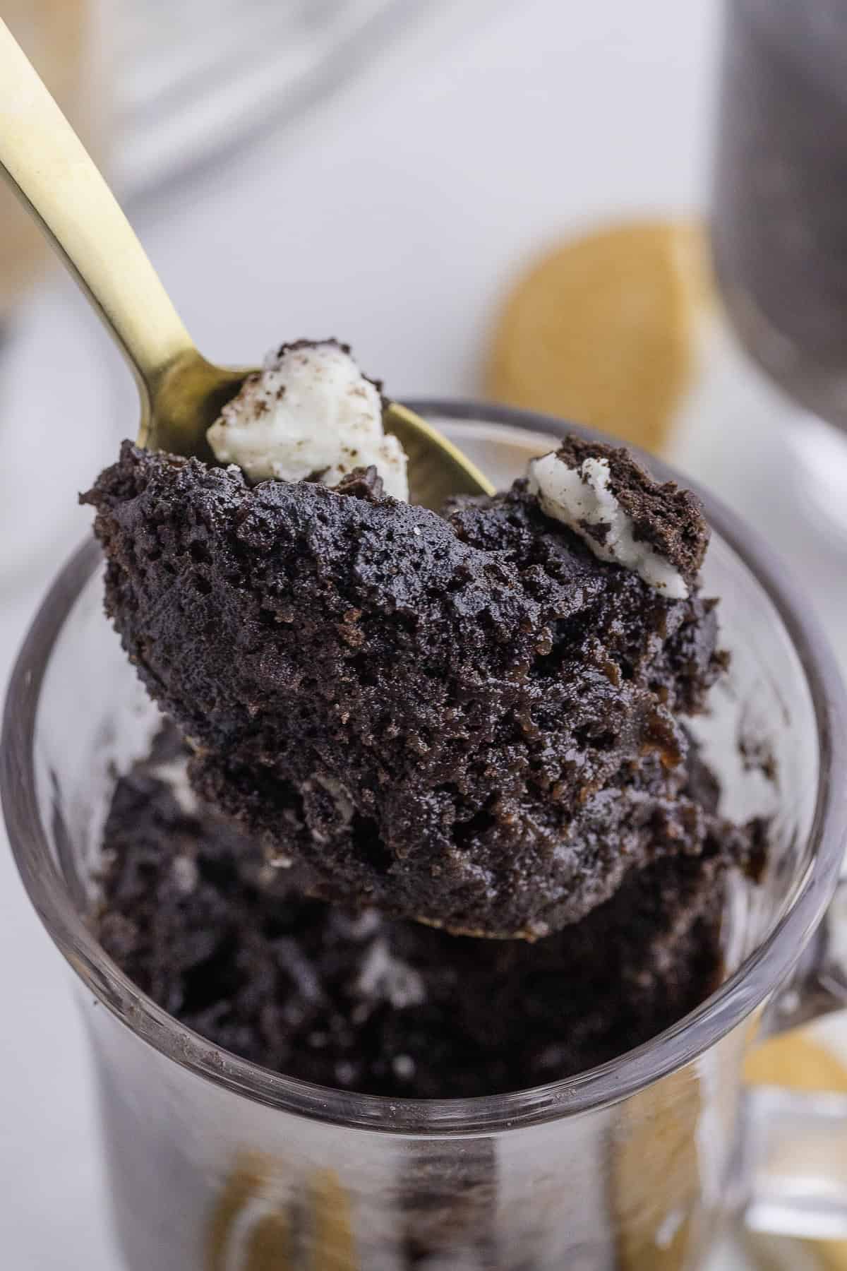 A spoon scoops chocolate mug cake topped with cream from a glass mug, with biscuits in the background.