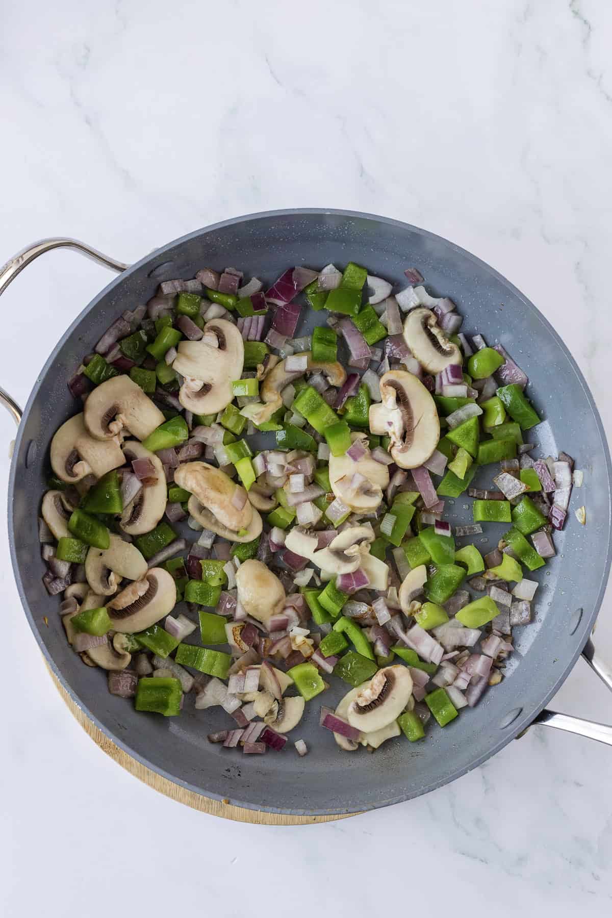 Sliced mushrooms, green bell peppers, and diced red onions cooking in a frying pan on a marble countertop.