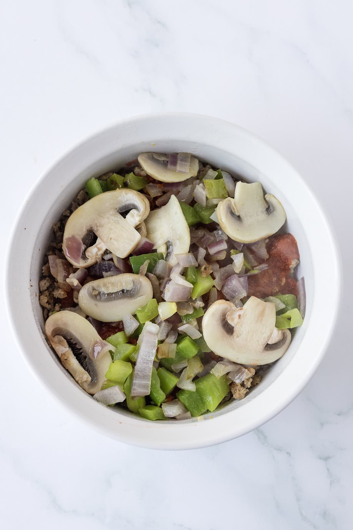 A bowl filled with sliced raw mushrooms, chopped green bell peppers, diced tomatoes, and onions.