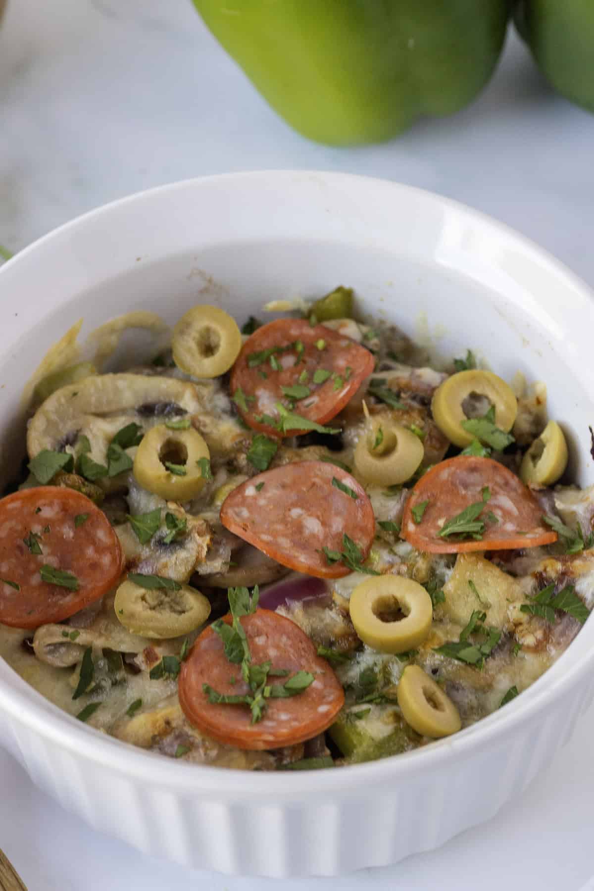 A white dish filled with a baked casserole topped with sliced pepperoni, green olives, and chopped herbs.