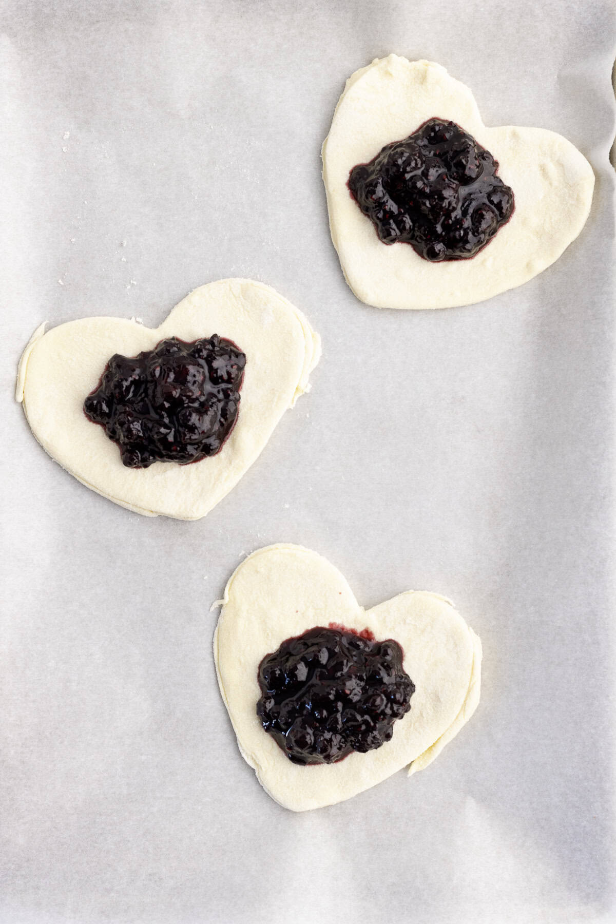 Three heart-shaped pieces of dough topped with dark fruit jam are arranged on a parchment-lined baking sheet.