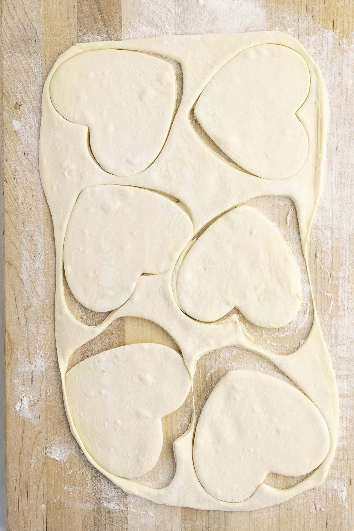 Heart-shaped pieces of dough cut from a rolled sheet on a floured wooden surface.