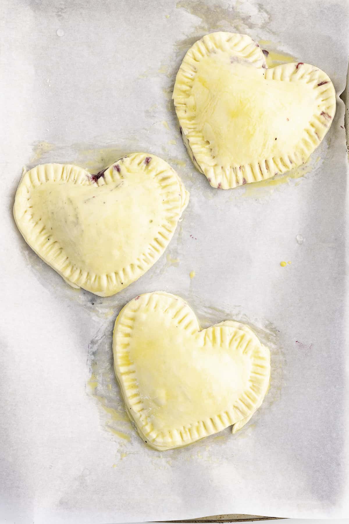 Three unbaked, heart-shaped hand pies with crimped edges are arranged on a parchment-lined baking sheet.