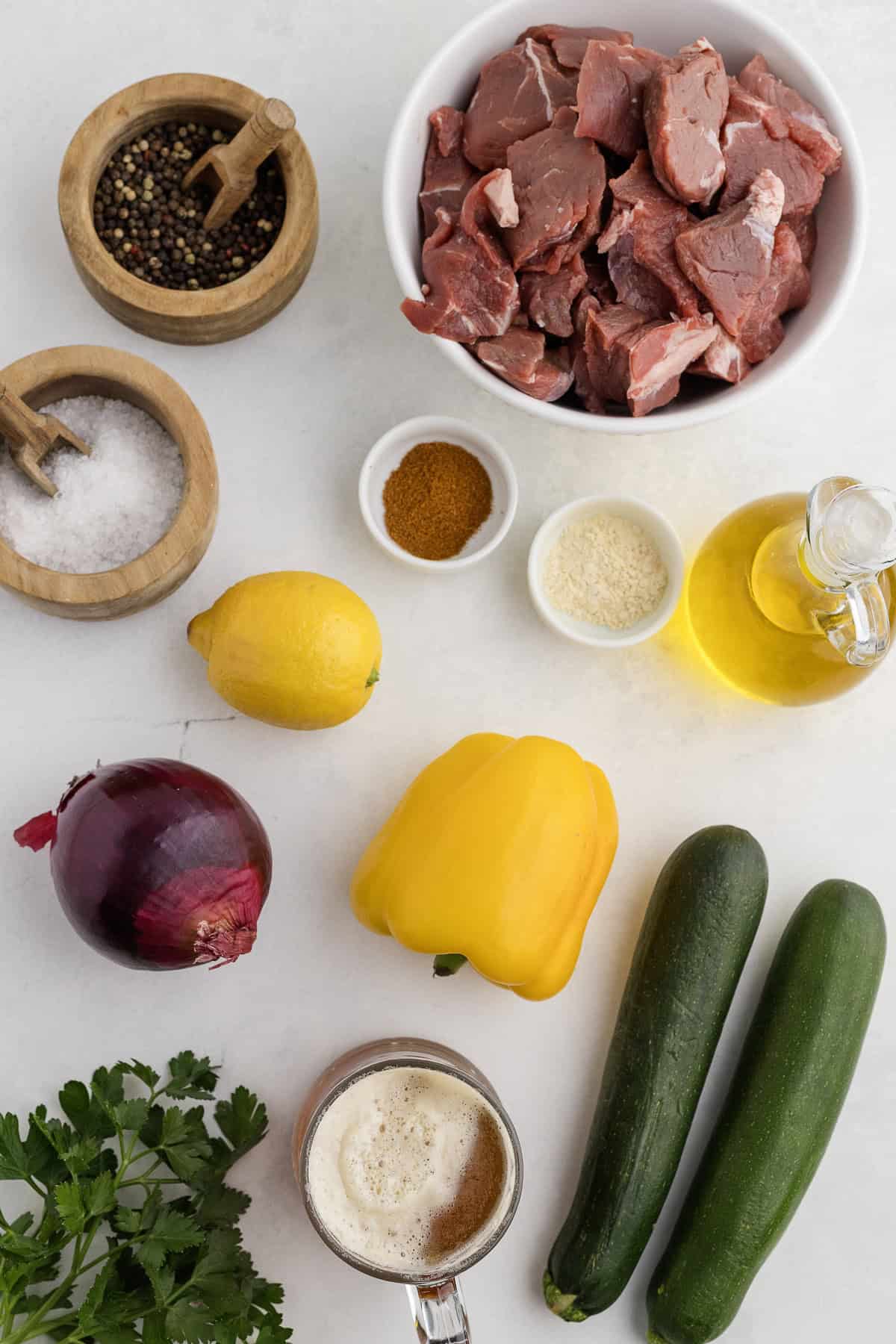 Assorted ingredients on a white surface: bowl of meat chunks, spices, oil, onion, yellow bell pepper, zucchini, lemons, parsley, salt, pepper, and a mug of frothy beer.