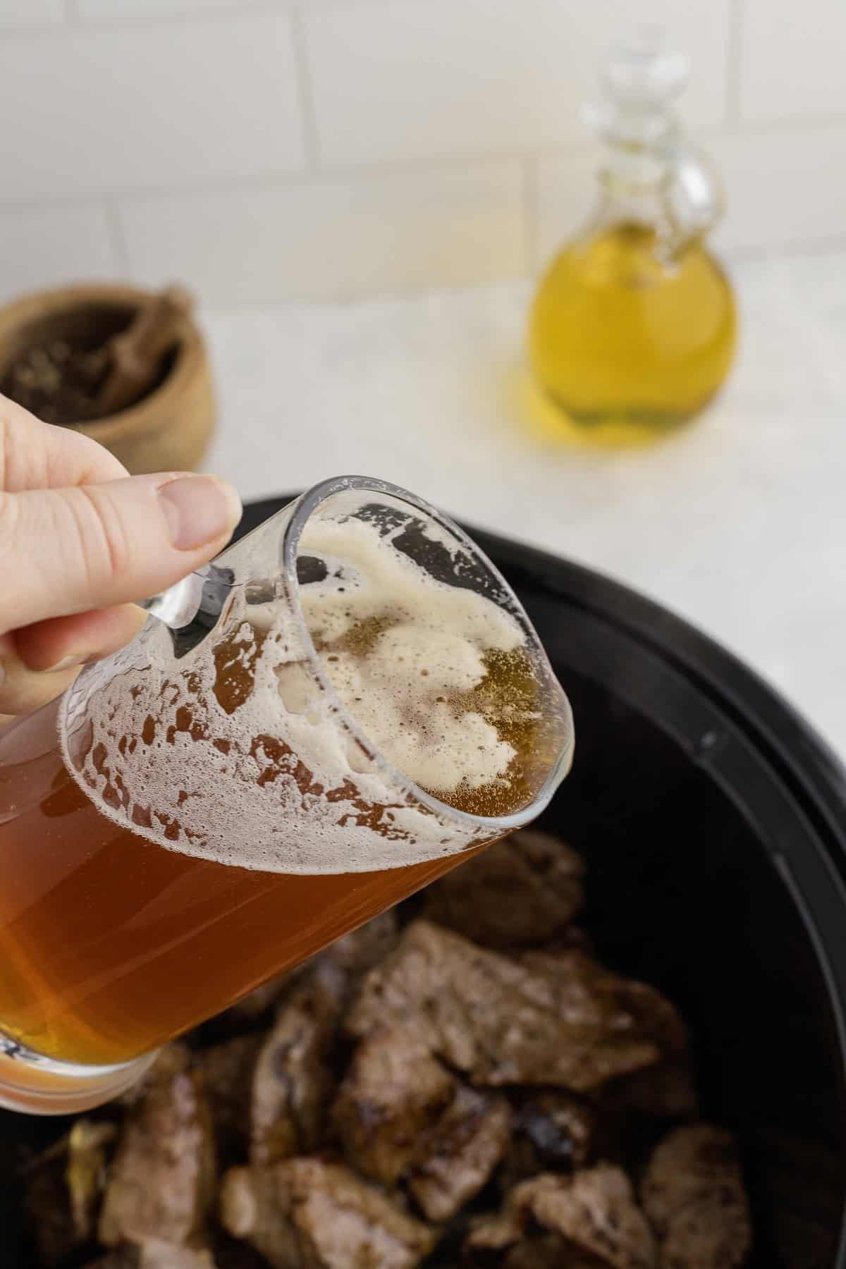 Person pouring beer from a glass into a slow cooker with browned meat, a small oil bottle in the background.