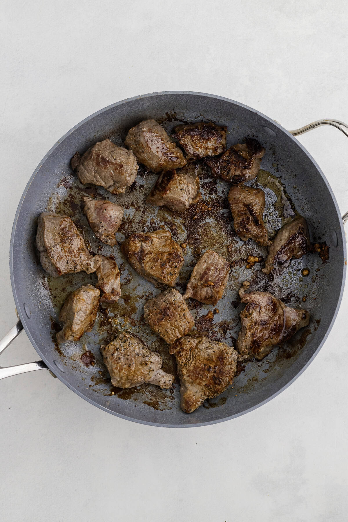 Chunks of browned meat cooking in a gray frying pan on a light surface.