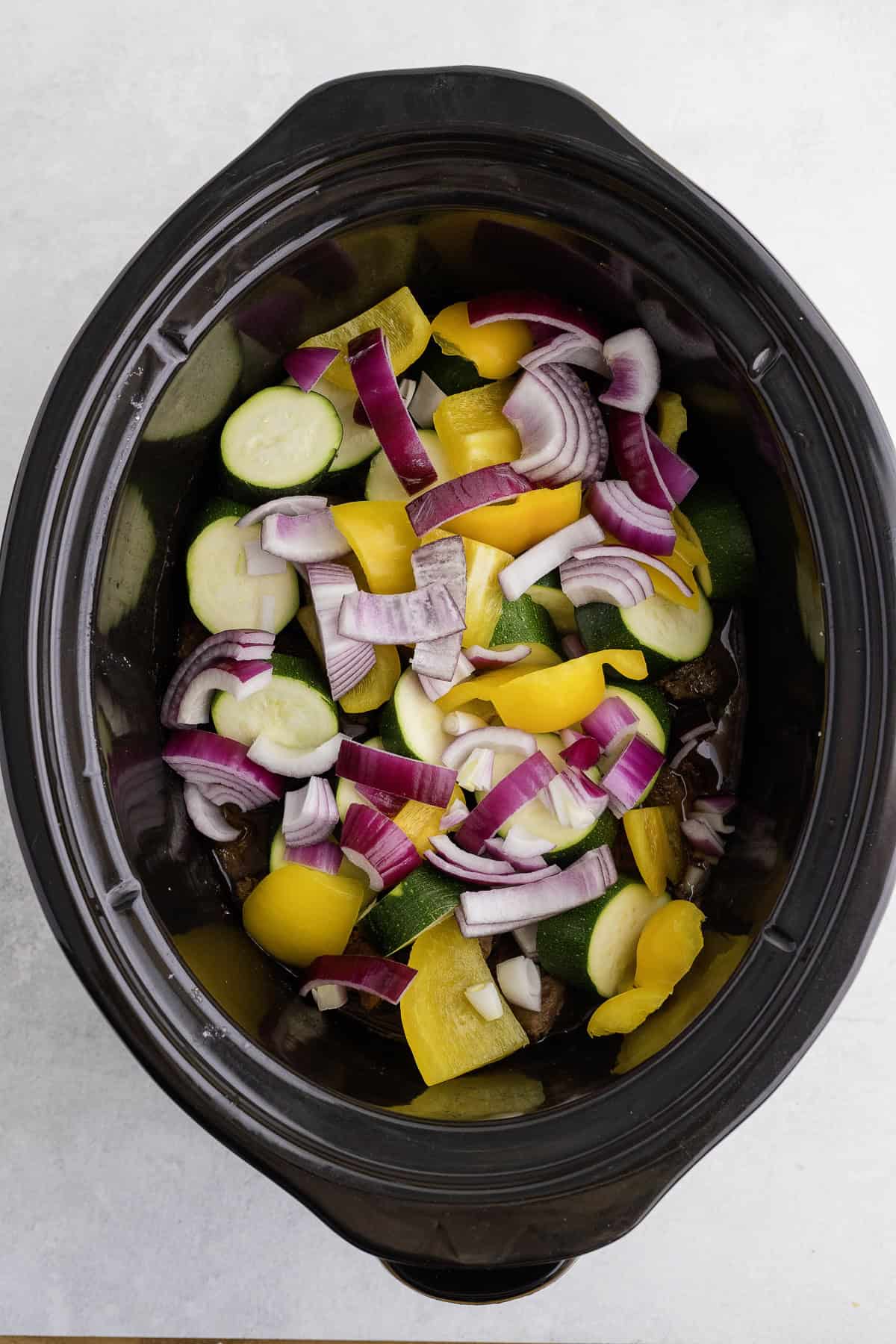 Chopped zucchini, yellow bell peppers, and red onions in a black slow cooker on a light surface.