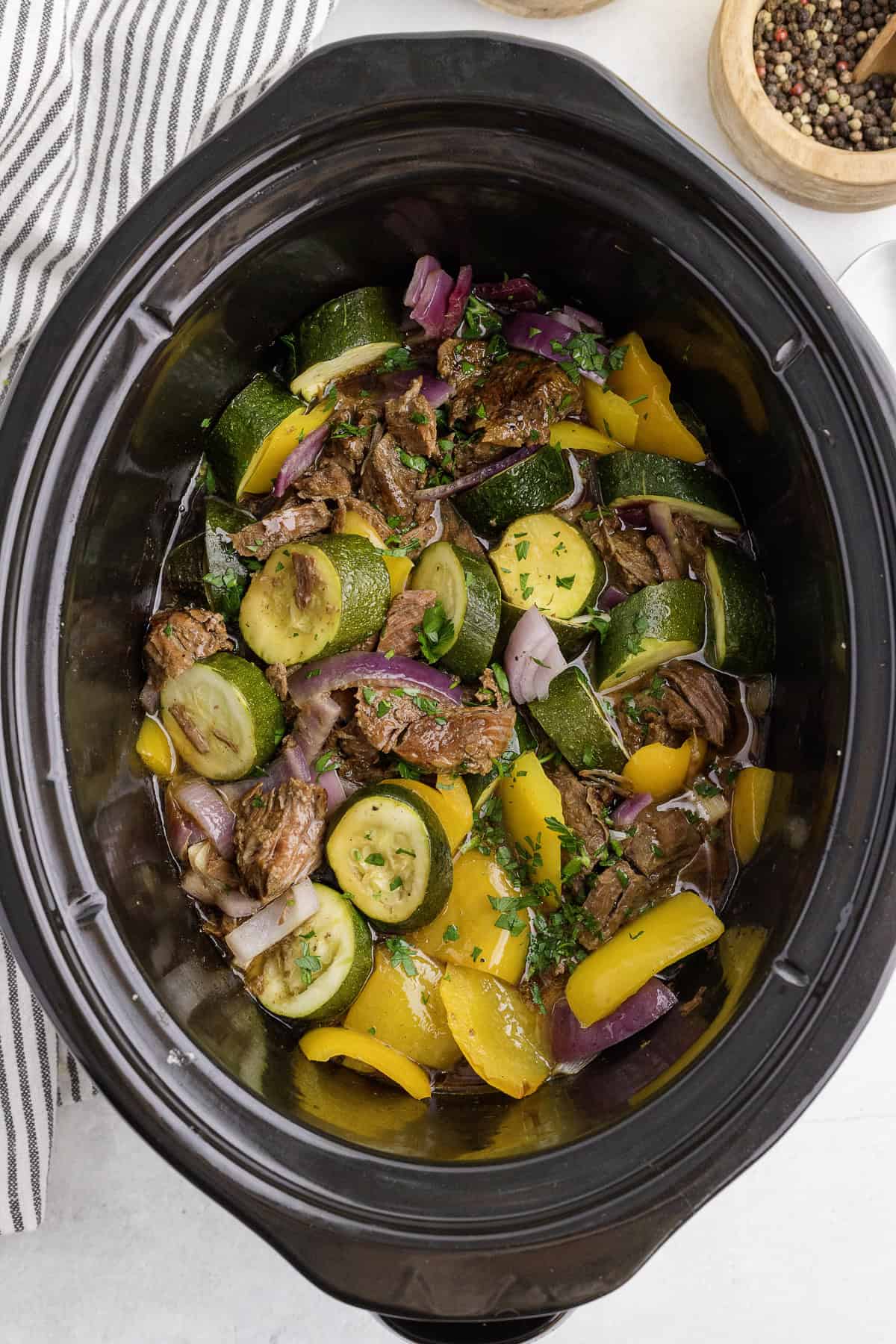 A slow cooker filled with beef chunks, zucchini slices, yellow bell peppers, and red onions, garnished with herbs.