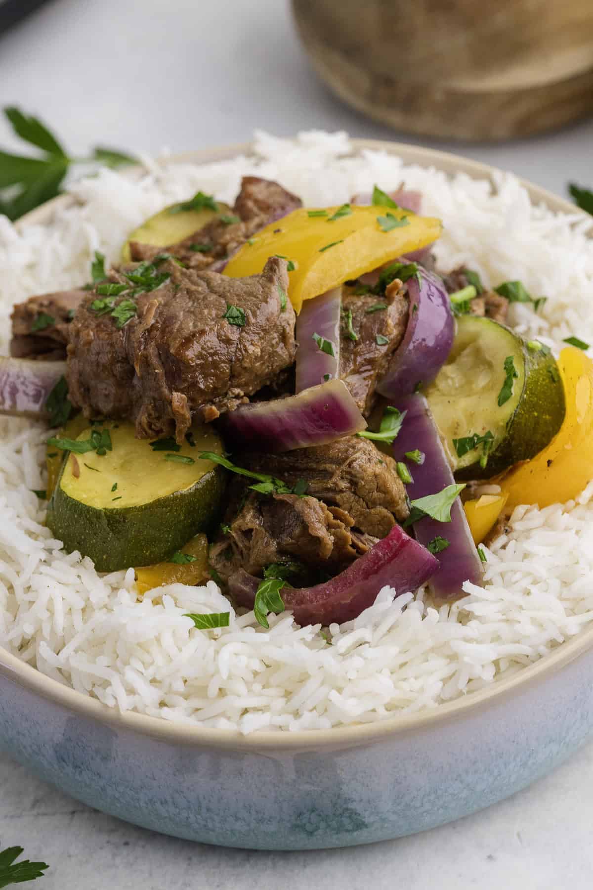 A bowl of white rice topped with pieces of beef, zucchini, yellow bell pepper, and red onion, garnished with chopped herbs.