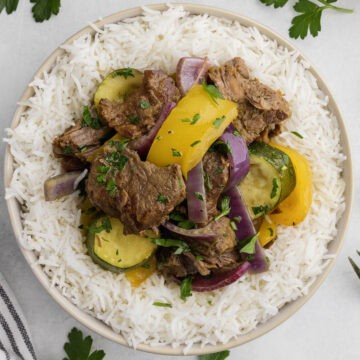 A bowl of rice topped with beef chunks, yellow bell pepper, zucchini, red onion, and parsley.