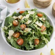 A bowl of mixed green salad with spinach, arugula, cherry tomatoes, and cheese slices, with vinaigrette in a small bowl beside it.