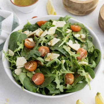 A bowl of mixed green salad with spinach, arugula, cherry tomatoes, and cheese slices, with vinaigrette in a small bowl beside it.