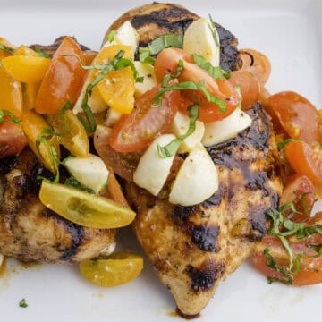 Grilled chicken breast topped with sliced cherry tomatoes, mozzarella pieces, and fresh basil on a white plate.