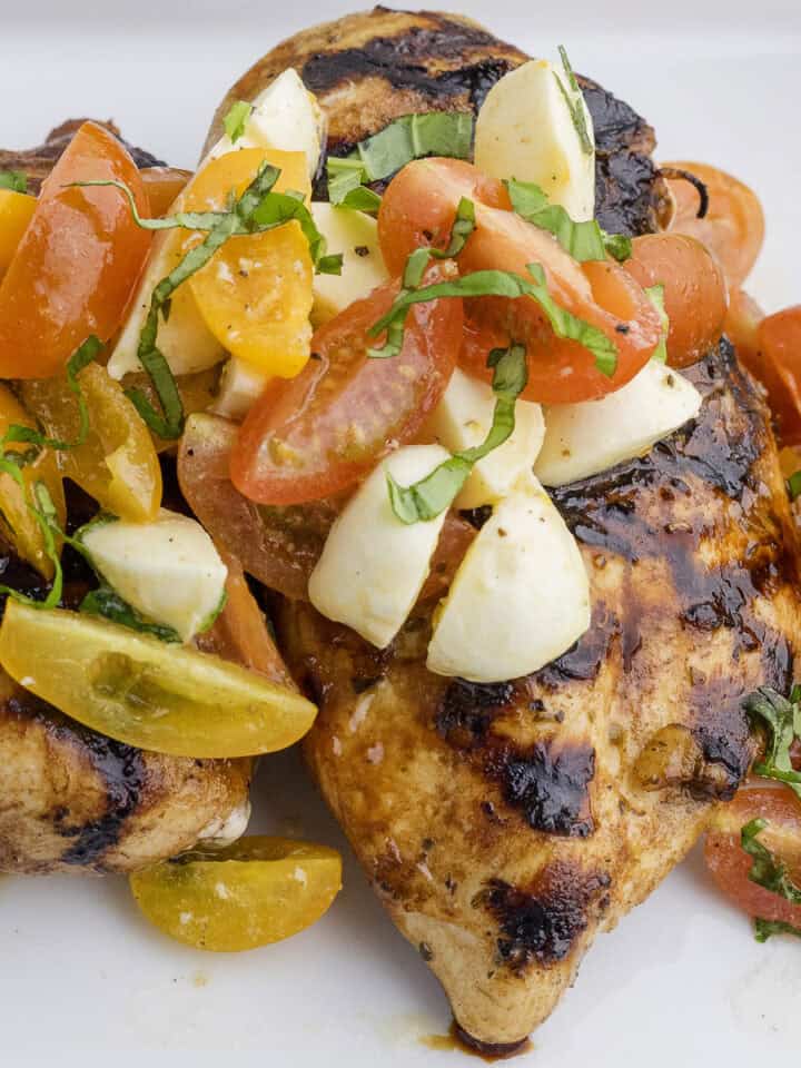 Grilled chicken breast topped with sliced cherry tomatoes, mozzarella pieces, and fresh basil on a white plate.