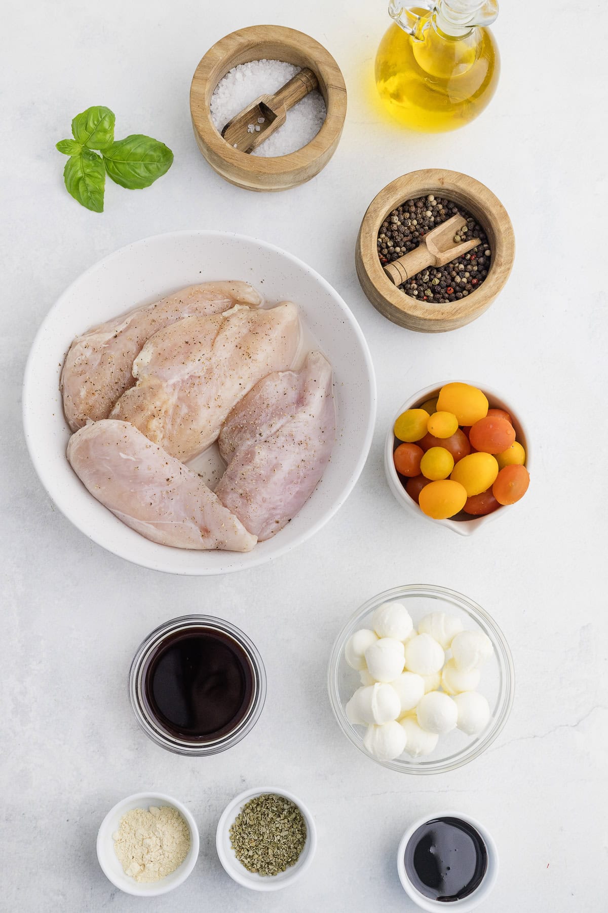 Raw chicken breasts in a bowl, surrounded by cherry tomatoes, mozzarella balls, olive oil, salt, pepper, balsamic vinegar, grated cheese, dried herbs, and fresh basil on a white surface.