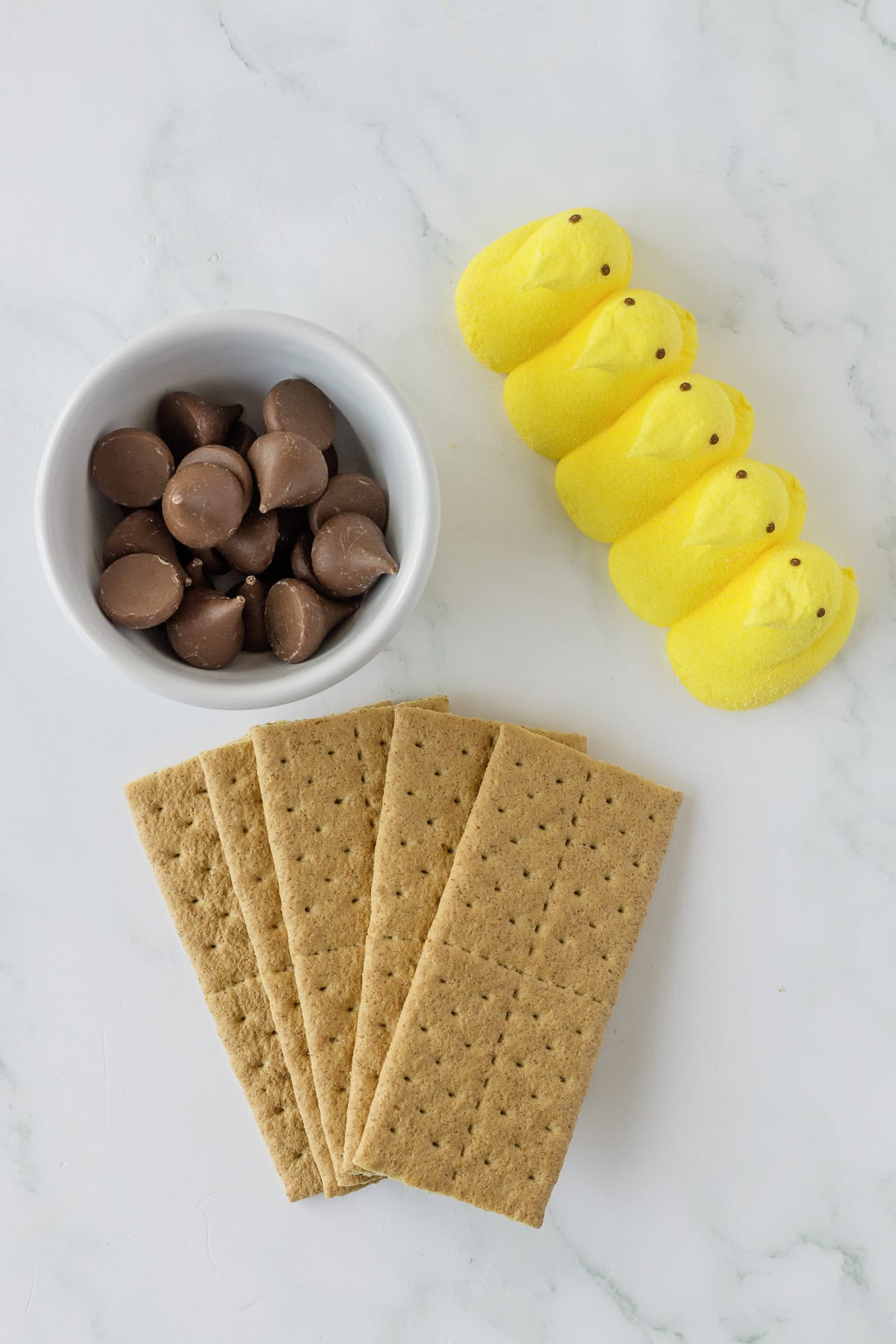 Graham crackers, chocolate candies in a bowl, and five yellow marshmallow chicks on a marble surface.