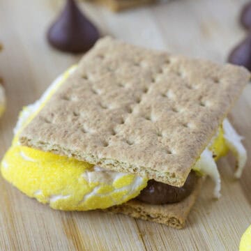 A s'more with a yellow marshmallow and chocolate between graham crackers on a wooden surface.
