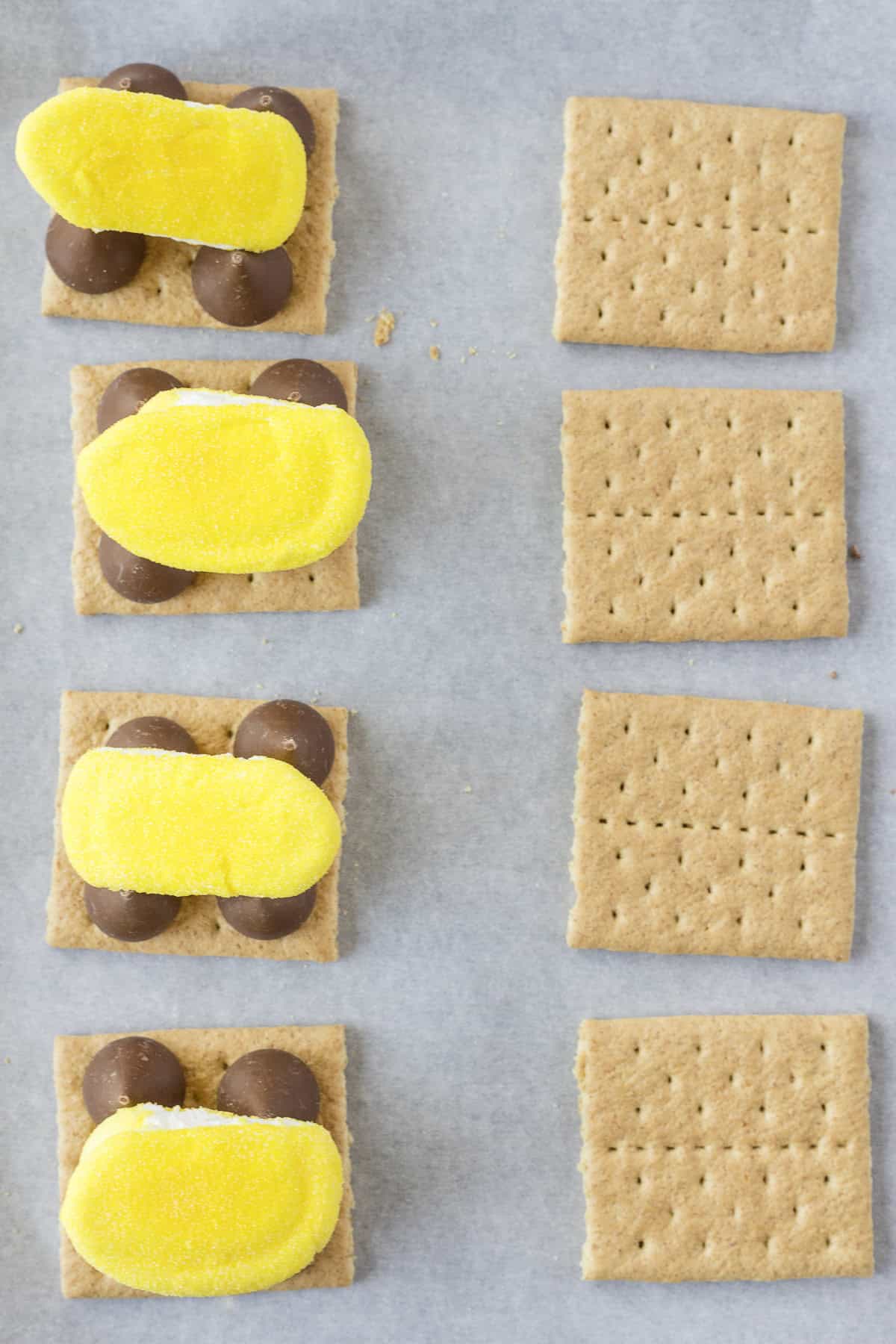 Graham crackers with yellow marshmallow chicks and chocolate pieces on the left; plain graham crackers on the right, on parchment paper.