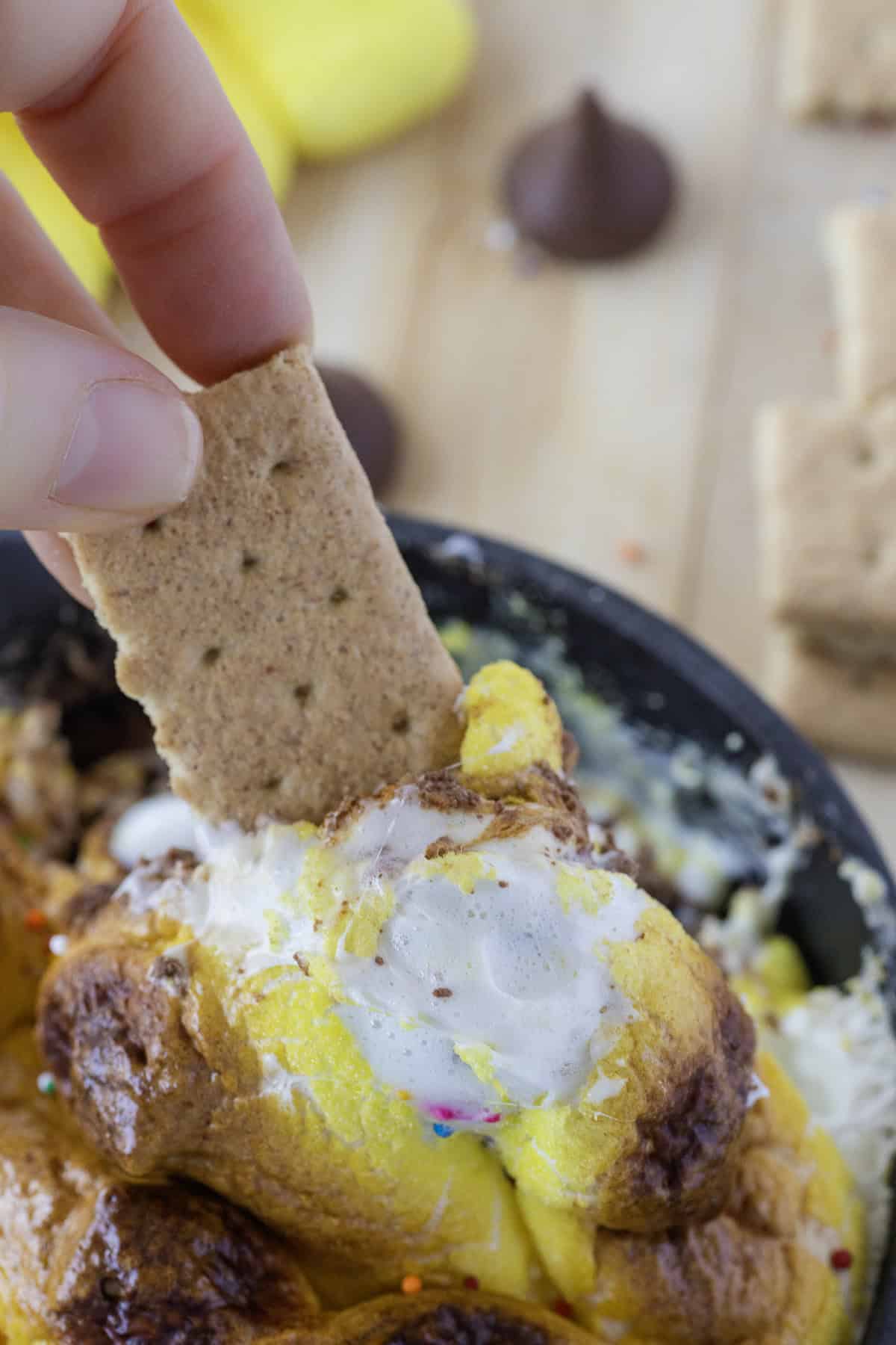Graham cracker dipped into melted marshmallow and chocolate in a bowl, with graham crackers and chocolate pieces in the background.