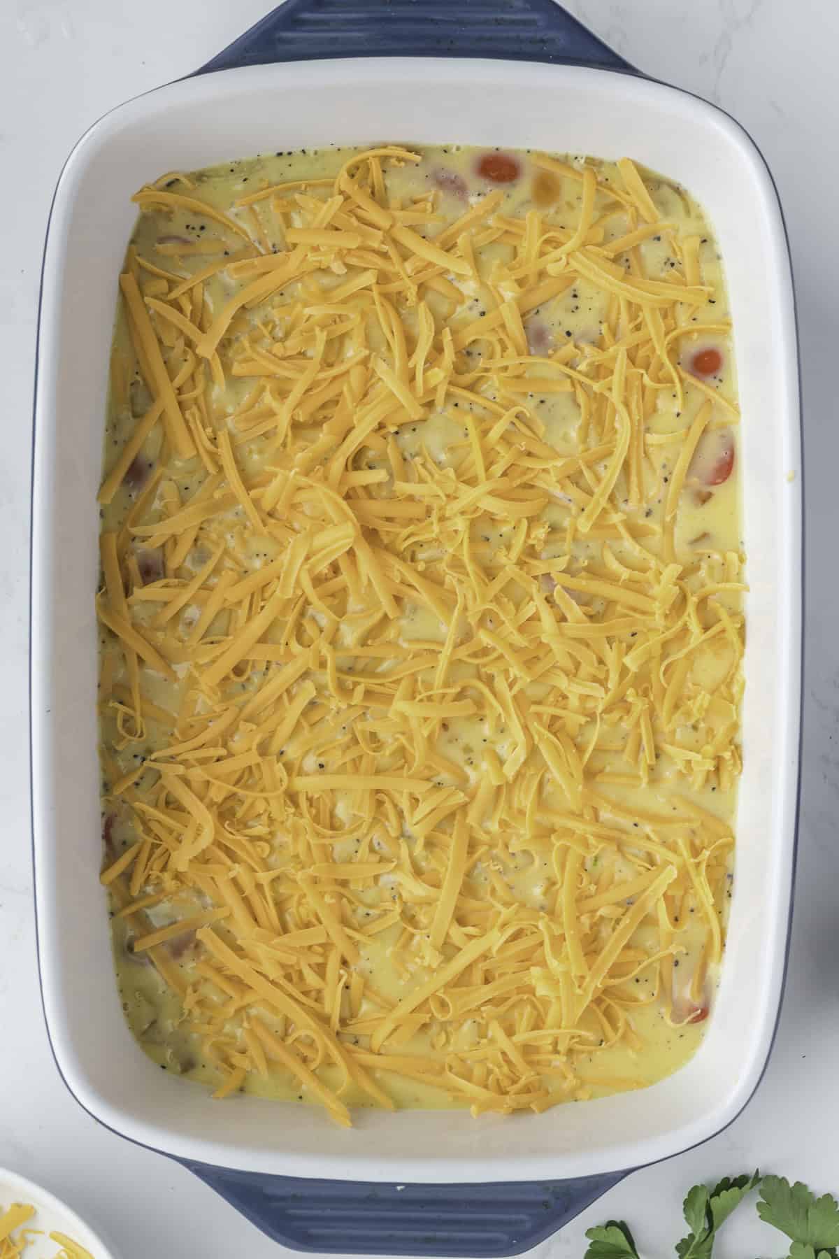 A rectangular baking dish filled with an unbaked casserole mixture, topped with shredded cheddar cheese.