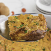 A close-up of a slice of baked egg casserole with herbs, held on a spatula above the remaining casserole in a baking dish.