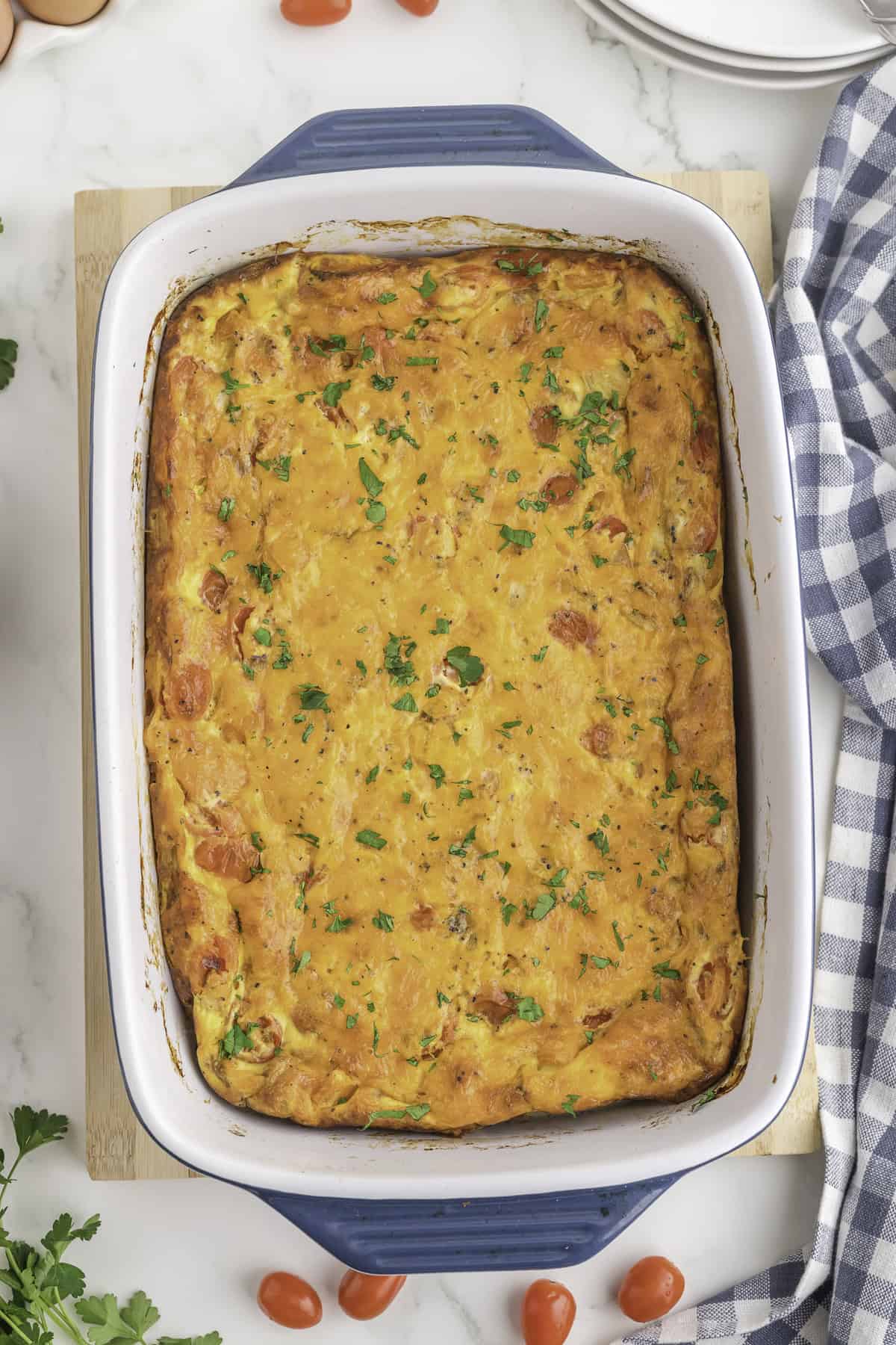 A baked casserole topped with chopped parsley in a white rectangular dish with blue handles, set on a wooden board beside a checkered cloth.