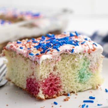 A slice of white cake with red and blue patches, topped with white frosting and blue and red sprinkles, sits on a white plate.