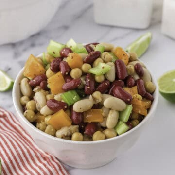 A white bowl filled with a mixed bean salad containing kidney beans, chickpeas, celery, yellow bell pepper, and white beans, set on a marble surface with lime wedges nearby.