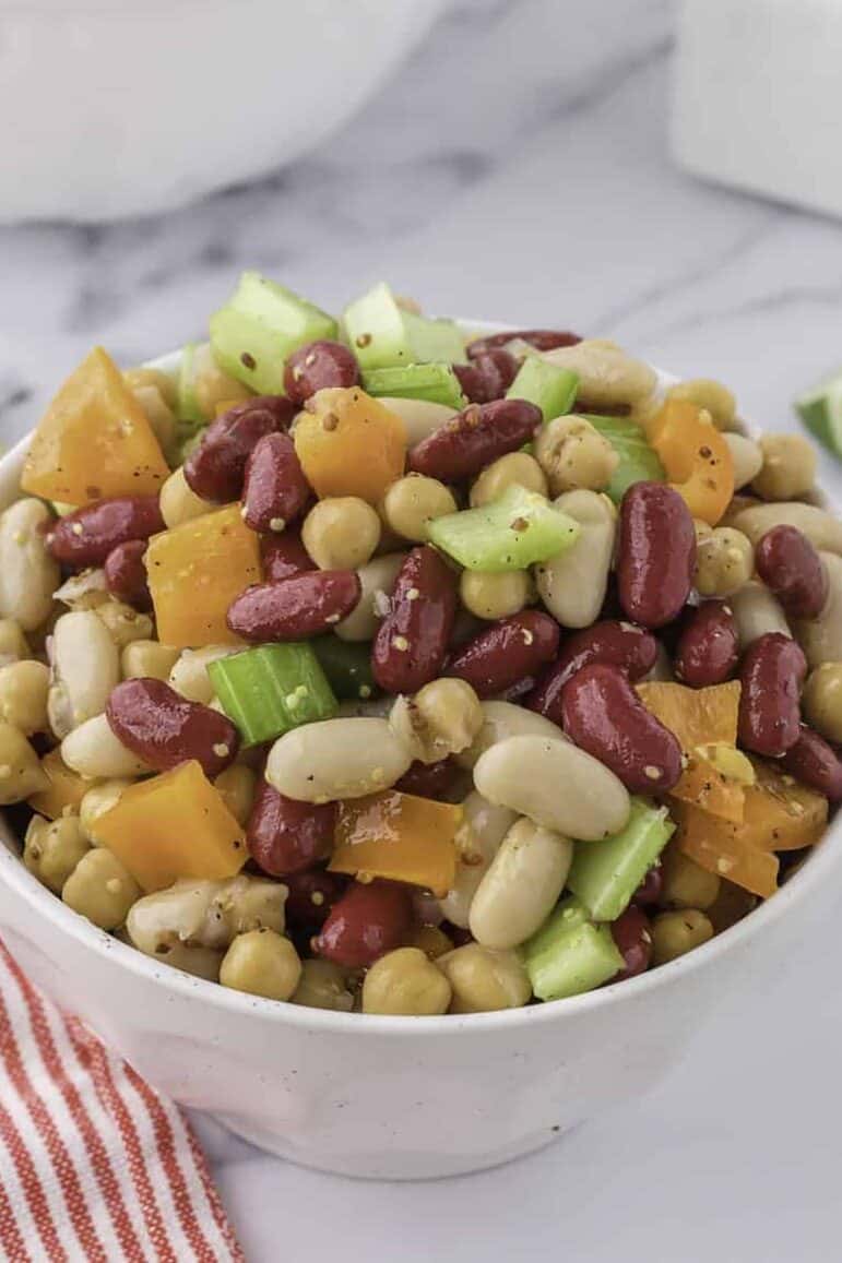 bowl of mixed bean salad with kidney beans, white beans, celery, and yellow bell peppers on a marble surface, with lime wedges.
